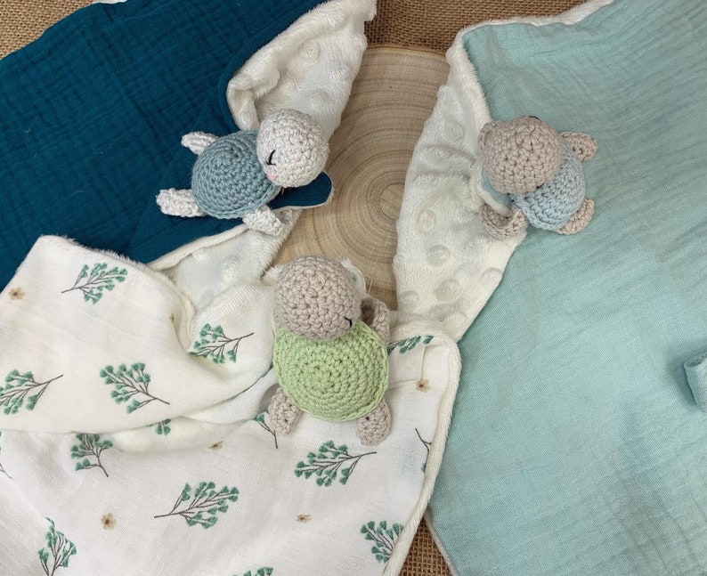 May include: Three crocheted turtle toys on a white, green, and blue fabric background. The turtles are different colors and have different colored shells.
