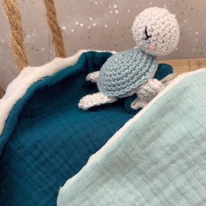 May include: A crocheted sea turtle toy with a light blue shell and white body, resting on a teal blue and light blue baby blanket with a white trim.
