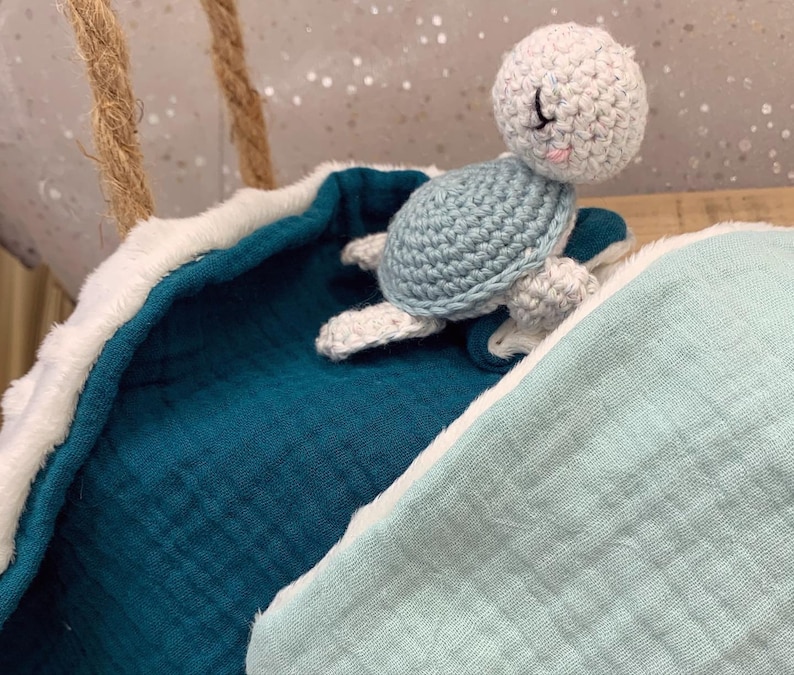 May include: A crocheted sea turtle toy with a light blue shell and white body, resting on a teal blue and light blue baby blanket with a white trim.