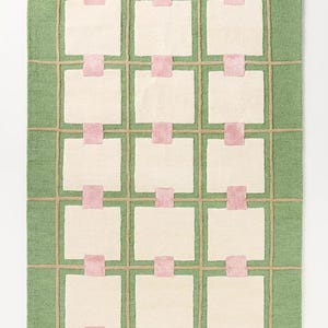 Pode incluir: Um tapete retangular com fundo verde e um padrão de grade. A grade é feita de linhas bege e emoldura quadrados brancos. Cada quadrado é conectado por um retângulo rosa. O tapete é um item decorativo para casa.