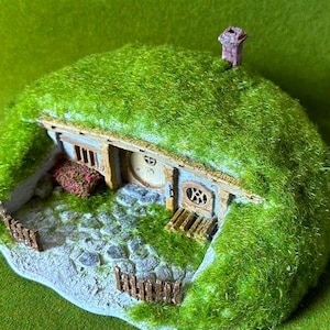 May include: A miniature hobbit house model with a green, grassy roof and a stone pathway leading to a round wooden door. The house has small windows and a chimney. The model is set against a green background.