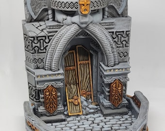 May include: A gray and gold miniature building with a stone archway and a sculpted face with a horned helmet. The building has a stone base with a decorative border and a small platform in front of the archway.