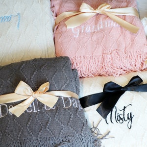 May include: Four knitted blankets with embroidered names and tied with satin ribbons. The blankets are in white, pink, gray, and white. The names on the blankets are "Grandma", "Misty", "Darrel & Darla", and "Laura".
