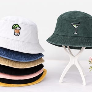 Könnte beinhalten: Eine Auswahl an bunten Fischerhüten, darunter Weiß, Jeansblau, Beige, Schwarz, Rosa und Gelb. Ein grüner Hut wird auf einem weißen Ständer präsentiert. Jeder Hut hat ein gesticktes Cocktail-Design, ideal für den Sommer.