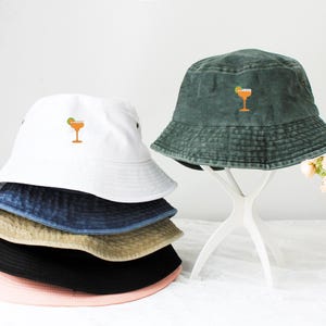Könnte beinhalten: Ein Stapel aus fünf Bucket-Hats in verschiedenen Farben, darunter rosa, schwarz, beige, blau und weiß. Der oberste Hut ist weiß mit einem gestickten orangen Cocktailglas-Design.