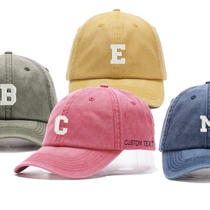 May include: Three baseball caps in different colours: olive green with a white "B", pink with a white "C", and blue with a white "M".