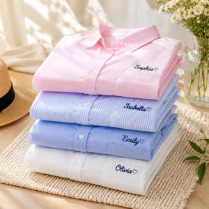 May include: A stack of four long-sleeved button-down shirts in pink, light blue, medium blue, and white. Each shirt features a chest pocket embroidered with a name and a small heart: Sophia, Isabella, Emily, and Olivia in dark blue script.