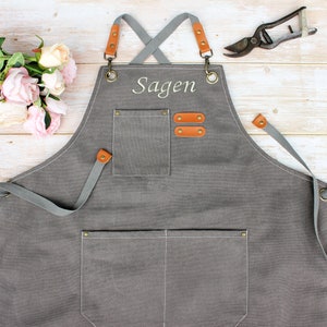 May include: A grey canvas apron with adjustable straps, a large pocket, and two smaller pockets. The apron is personalised with the name "Sagen" in white embroidery.