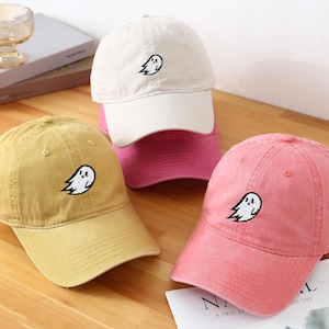 Puede incluir: Varias gorras de béisbol en tonos mostaza, crema y rosa. Cada gorra presenta un diseño de fantasma bordado. Las gorras están hechas de una tela suave y texturizada y tienen una visera curva. Un accesorio de moda.