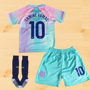 以下が含まれることがあります： サッカーユニフォームセット。ジャージ、ショーツ、ソックスが含まれています。ジャージは水色、ピンク、紫で、背中には「LAMINE YAMAL」と数字の「10」が入っています。ショーツは水色で数字の「10」が入っています。ネイビーブルーのソックスには「BARÇA」の文字が入っています。