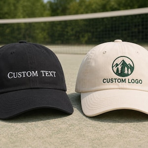 Può includere: Un cappellino da baseball nero con testo ricamato in bianco CUSTOM TEXT e un cappellino da baseball color crema con un grafico circolare ricamato in verde di montagne e pini sopra il testo verde ricamato CUSTOM LOGO.