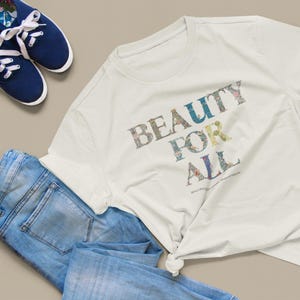 A cream t-shirt with "BEAUTY FOR ALL" in floral print is tied at the bottom, next to a pair of blue jeans and navy sneakers.