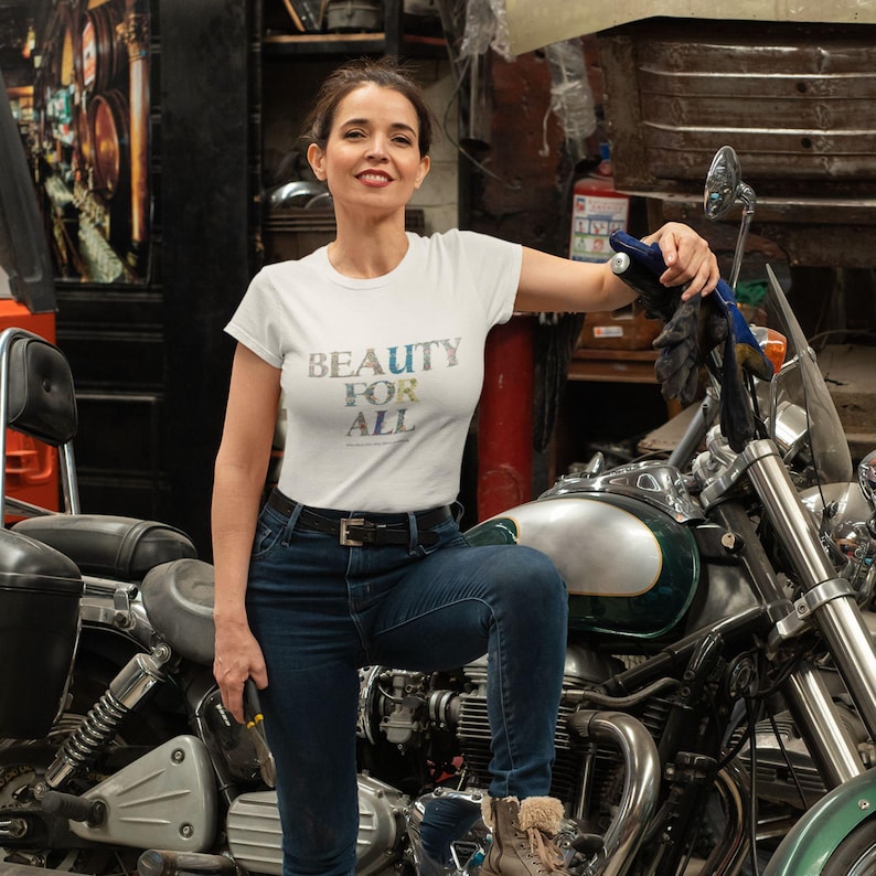 A woman with dark hair and a white t-shirt with "BEAUTY FOR ALL" in floral print stands next to a motorcycle in a garage.