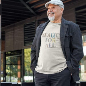 A man with a white beard and a gray cap wears a cream t-shirt with "BEAUTY FOR ALL" in floral print under a dark jacket.