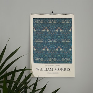 A William Morris 2025 wall calendar hanging on a light grey wall with a single bird pattern in various shades of blue, alongside text at the bottom.