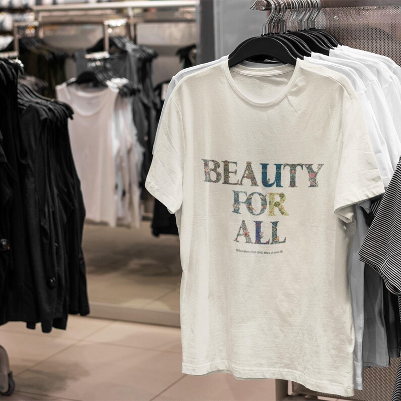 A cream t-shirt with "BEAUTY FOR ALL" in floral print hangs on a rack with other white shirts in a store setting.