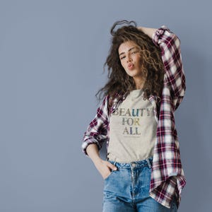 A woman with curly hair wearing a plaid shirt over a cream t-shirt with "BEAUTY FOR ALL" in floral print, making a kissy face.
