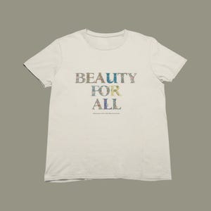 A cream-colored short-sleeved t-shirt with the words "BEAUTY FOR ALL" printed on the front in floral and nature-themed patterns, with a small quote below.