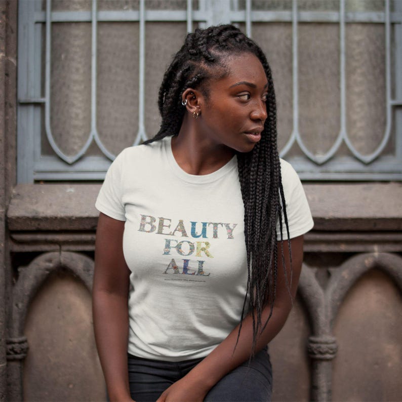 A woman with long dark braids wears a cream t-shirt with "BEAUTY FOR ALL" in floral print, looking to her right against a stone wall.