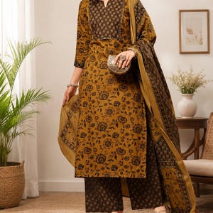 May include: A mustard yellow and dark brown three-piece outfit consisting of a long-sleeved tunic with a notch neckline, wide-leg trousers, and a matching dupatta. The fabric features a repeating floral print with dark brown patterns throughout.