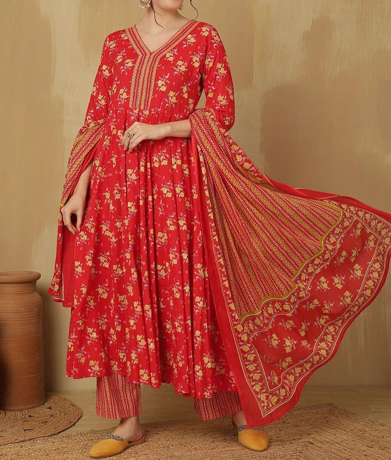 May include: A red floral print ensemble featuring a long-sleeved top with a V-neck and matching pants. The outfit includes a flowing scarf with a striped and floral pattern. The fabric is adorned with yellow floral designs.