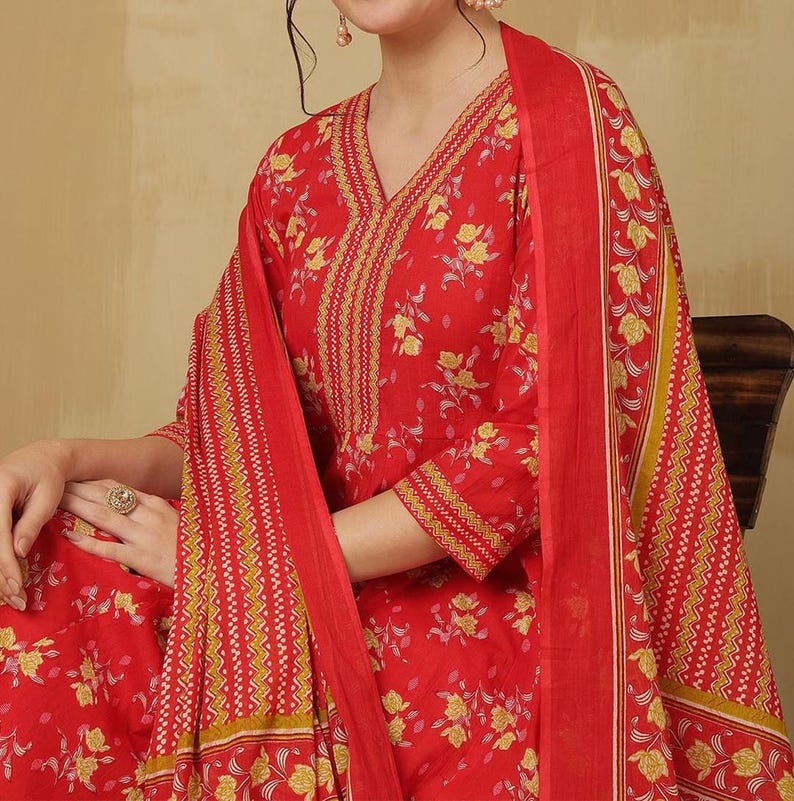 May include: A red floral print salwar kameez set. The outfit features a V-neck top with three-quarter sleeves and a matching scarf. The fabric is adorned with yellow floral patterns and intricate border designs.