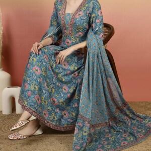 May include: A teal floral print dress with a matching shawl. The dress features a V-neckline and three-quarter sleeves. The floral pattern includes pink and yellow flowers with green leaves. The shawl is draped over the chair and has a similar floral design.