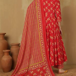 May include: A red floral print Anarkali suit with a matching dupatta. The suit features a three-quarter sleeve top and matching pants. The dupatta has a striped pattern with floral borders. The outfit is made of a lightweight fabric.