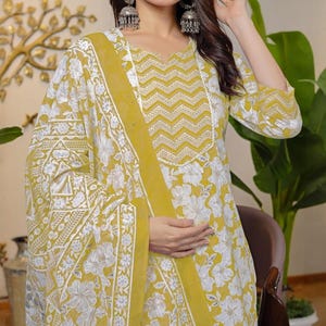 Puede incluir: Un conjunto amarillo con estampado floral, flores blancas y bordados intrincados. El conjunto incluye una blusa de manga larga con un diseño de chevrón en el escote y una bufanda a juego. La tela es ligera.