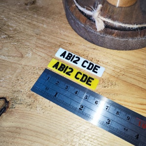 May include: Two rectangular stickers with the text "ABI2 CDE" in black, one white and one yellow. A blue ruler with inch measurements is next to the stickers. The items are on a wooden surface.