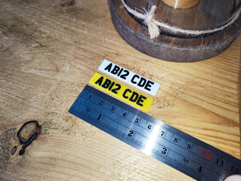 May include: Two rectangular stickers with the text "ABI2 CDE" in black, one white and one yellow. A blue ruler with inch measurements is next to the stickers. The items are on a wooden surface.