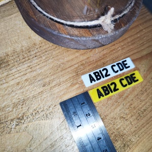 May include: Two rectangular stickers with the text "ABI2 CDE" in black. One sticker is white, the other is yellow. A metal ruler with inch measurements is next to the stickers. A wooden bowl with a rope trim is in the background.