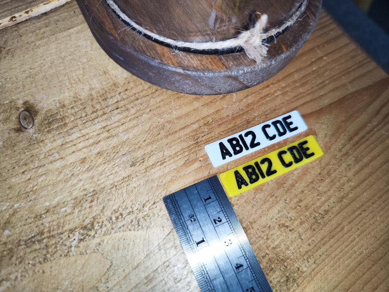 May include: Two rectangular stickers with the text "ABI2 CDE" in black. One sticker is white, the other is yellow. A metal ruler with inch measurements is next to the stickers. A wooden bowl with a rope trim is in the background.