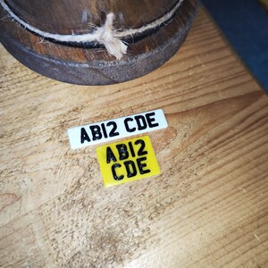 May include: Two small rectangular license plates with the text "ABI2 CDE" in black. One plate is white, the other is yellow. A wooden object with a rope is in the background. The items are on a wooden surface.