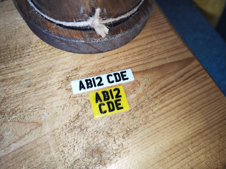 May include: Two small rectangular license plates with the text "ABI2 CDE" in black. One plate is white, the other is yellow. A wooden object with a rope is in the background. The items are on a wooden surface.