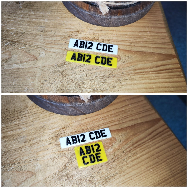 May include: Two small rectangular license plates, one white and one yellow, each with the text "ABI2 CDE" in black. The plates are on a wooden surface, with a dark brown circular object and rope in the background.