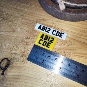 May include: Two small rectangular plates with the text "ABI2 CDE" in black. One plate is white, the other is yellow. A metal ruler with inch markings is also present, measuring up to 6 inches.