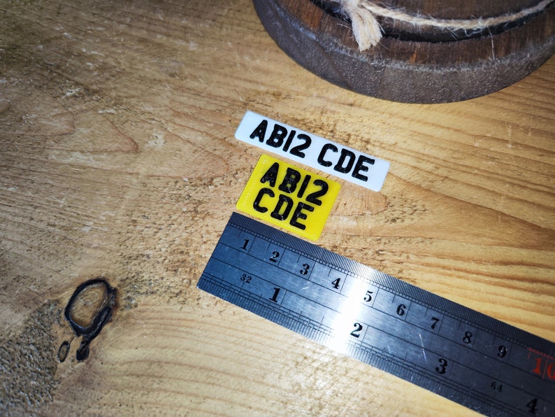 May include: Two small rectangular plates with the text "ABI2 CDE" in black. One plate is white, the other is yellow. A metal ruler with inch markings is also present, measuring up to 6 inches.