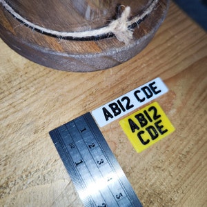 May include: Two small rectangular license plates with the text "ABI2 CDE" in black. One plate is white, the other is yellow. A ruler with inch measurements is next to the plates. A wooden bowl is in the background.