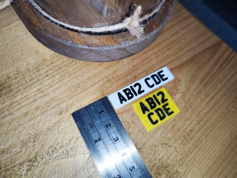 May include: Two small rectangular license plates with the text "ABI2 CDE" in black. One plate is white, the other is yellow. A ruler with inch measurements is next to the plates. A wooden bowl is in the background.