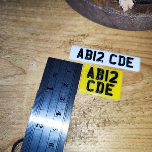 May include: Two miniature license plates, one white and one yellow, each displaying the text "ABI2 CDE" in black. A metal ruler with inch markings is positioned next to the plates on a wooden surface.