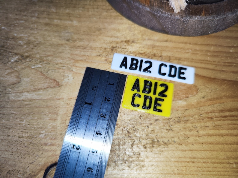 May include: Two miniature license plates, one white and one yellow, each displaying the text "ABI2 CDE" in black. A metal ruler with inch markings is positioned next to the plates on a wooden surface.
