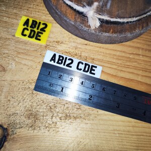 May include: Two rectangular labels with the text "ABI2 CDE" are displayed on a wooden surface. A metal ruler is placed over one label. A yellow label is also present. The image includes a wooden object with twine.