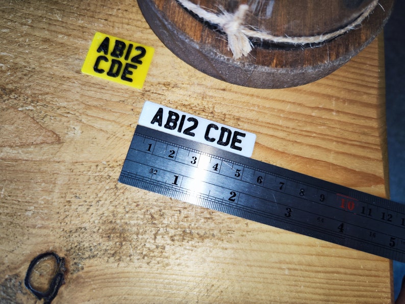 May include: Two rectangular labels with the text "ABI2 CDE" are displayed on a wooden surface. A metal ruler is placed over one label. A yellow label is also present. The image includes a wooden object with twine.