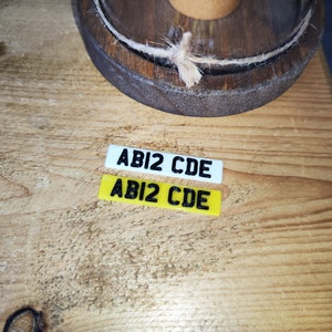 May include: Two small rectangular license plates with the text "ABI2 CDE" in black. One plate is white, the other is yellow. The plates are on a wooden surface, with a wooden barrel top in the background.
