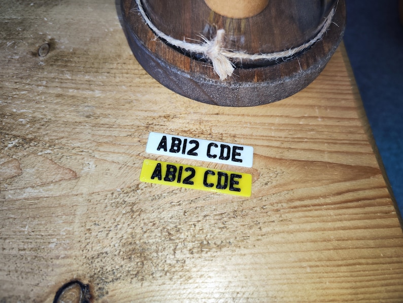 May include: Two small rectangular license plates with the text "ABI2 CDE" in black. One plate is white, the other is yellow. The plates are on a wooden surface, with a wooden barrel top in the background.