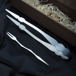 May include: A set of stainless steel serving utensils, including tongs and a fork, presented on a black surface. The utensils have a unique, wavy design. A wooden box with wood shavings is in the background.