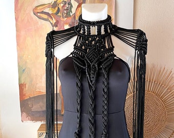 Peut inclure: Un ensemble noir en macramé pour les épaules et le cou, avec de longues franges et des détails tressés. L'accessoire fait main est présenté sur un mannequin portant une robe noire. L'arrière-plan comprend une peinture et des objets décoratifs.