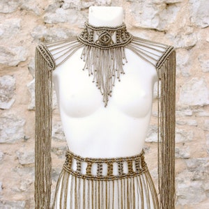 Op de afbeelding: Een gouden kralenketting en bijpassende riem met franjes. De ketting is een choker met een geometrisch ontwerp. De riem is een brede band met een vergelijkbaar geometrisch ontwerp.