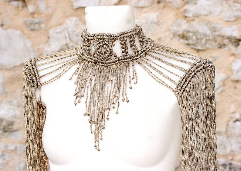 Op de afbeelding: Een gouden kralenketting met een franje-ontwerp. De ketting is gemaakt van kleine, ronde kralen die in een patroon aan elkaar geregen zijn. De franje is gemaakt van lange strengen kralen die van de ketting hangen. De ketting is een statement-stuk dat perfect zou zijn voor een speciale gelegenheid.